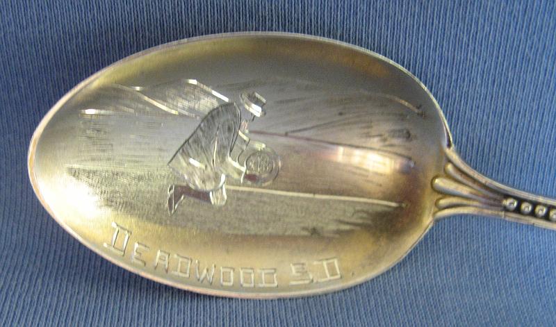 Souvenir Mining Spoon Bowl Deadwood SD.JPG - SOUVENIR MINING SPOON DEADWOOD SD - Sterling silver spoon with engraved bowl showing miner panning for gold and marked DEADWOOD S.D., figuralhandle with gold pan, pick, shovel and ore bucket on top with rope wrappingthe top of handle, 5 3/8 in. long, reverse marked Sterling with CB&H hallmark for Codding Brothers & Heilbron, North Attleboro, MA who made sterling flatware from 1879-1918, Heilbron became a partner in 1891 [Deadwood, South Dakota was established in 1876 during the Black Hills gold rush. The settlement of Deadwood began illegally on land which had been granted to the Lakota-Sioux.  In 1875, a miner named John B. Pearson found gold in a narrow canyon in the Northern Black Hills. This canyon became known as "Deadwood Gulch," because of the many dead trees that lined the canyon walls at the time. The name stuck.  Today Deadwood has a population of 1270 residents and serves as the county seat of Lawrence County.  In 1874 a government- sponsored expedition confirmed the presence of gold in the Black Hills. The U.S. government tried to conceal the discovery from the general public in order to honor the 1868 Treaty of Fort Laramie, which forever ceded the Black Hills to the Lakota-Sioux. Despite the efforts of the military and federal government, the American populace learned about the discovery of gold in the Black Hills. Influenced by dreams and greed, the 1876 gold rush was on in the Black Hills. Once Deadwood was established, the mining camp was soon swarming with thousands of prospectors searching for an easy way to get rich. Most of the early population was in Deadwood to mine for gold, but the lawless region naturally attracted a crowd of rough and shady characters. These particular individuals made the early days of Deadwood rough and wild. A mostly male population eagerly patronized the many saloons, gambling establishments, dance halls, and brothels. By late 1877, Deadwood was moving from a primitive mining camp to a community with a town government, wood and brick buildings, and a sense of law and order. As the economy changed from gold panning to deep mining, Deadwood lost its rough and rowdy character. The town survived two devastating fires, one in 1879 and another in 1894, each time to rebuild.  In 1890, the Fremont, Elkhorn, and Missouri Valley Railroad connected the town to the outside world.  With the railroad, Deadwood evolved into a prosperous commercial center primarily focused on its gold mining industry.  South Dakota entered statehood on November 2, 1889 and Deadwood became the place where people traveled in the Black Hills to conduct their business, settling into the twentieth century.  Deadwood became the only city in the United States to be named a National Historic Landmark in 1964. A state initiative authorized legalized gaming in Deadwood to start on November 1, 1989. The introduction of gaming has enabled Deadwood to preserve its historic buildings and dramatically increase tourism. The lure of gaming is not the only draw to Deadwood; people are also fascinated by its unique, colorful history.]